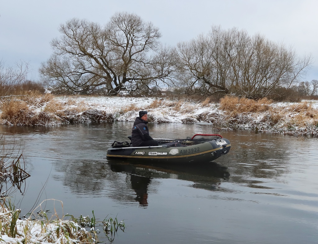 Рыбалка зимой с лодки Рыболов в надувной лодке на реке с заснеженными берегами и голым деревом на заднем плане