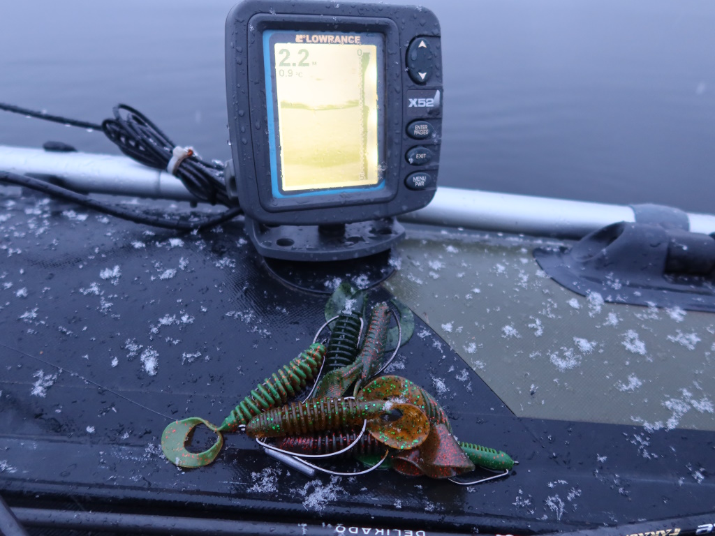 Эхолот Эхолот Lowrance на борту лодки показывает глубину и температуру воды, рядом лежат силиконовые приманки на заснеженном борту лодки
